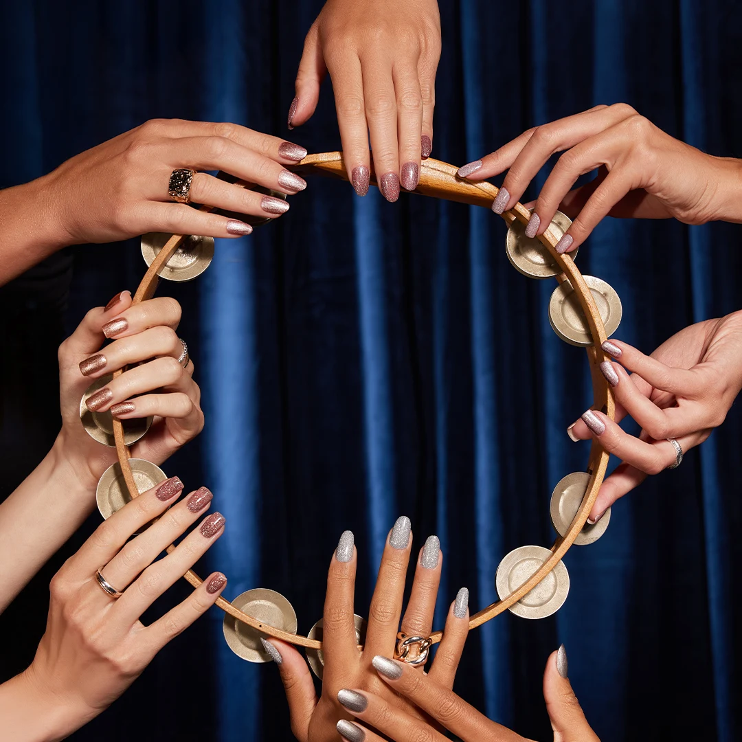 Wiele kobiecych dłoni z ozdobnymi paznokciami i biżuterią trzyma tamburyn na tle granatowej zasłony.