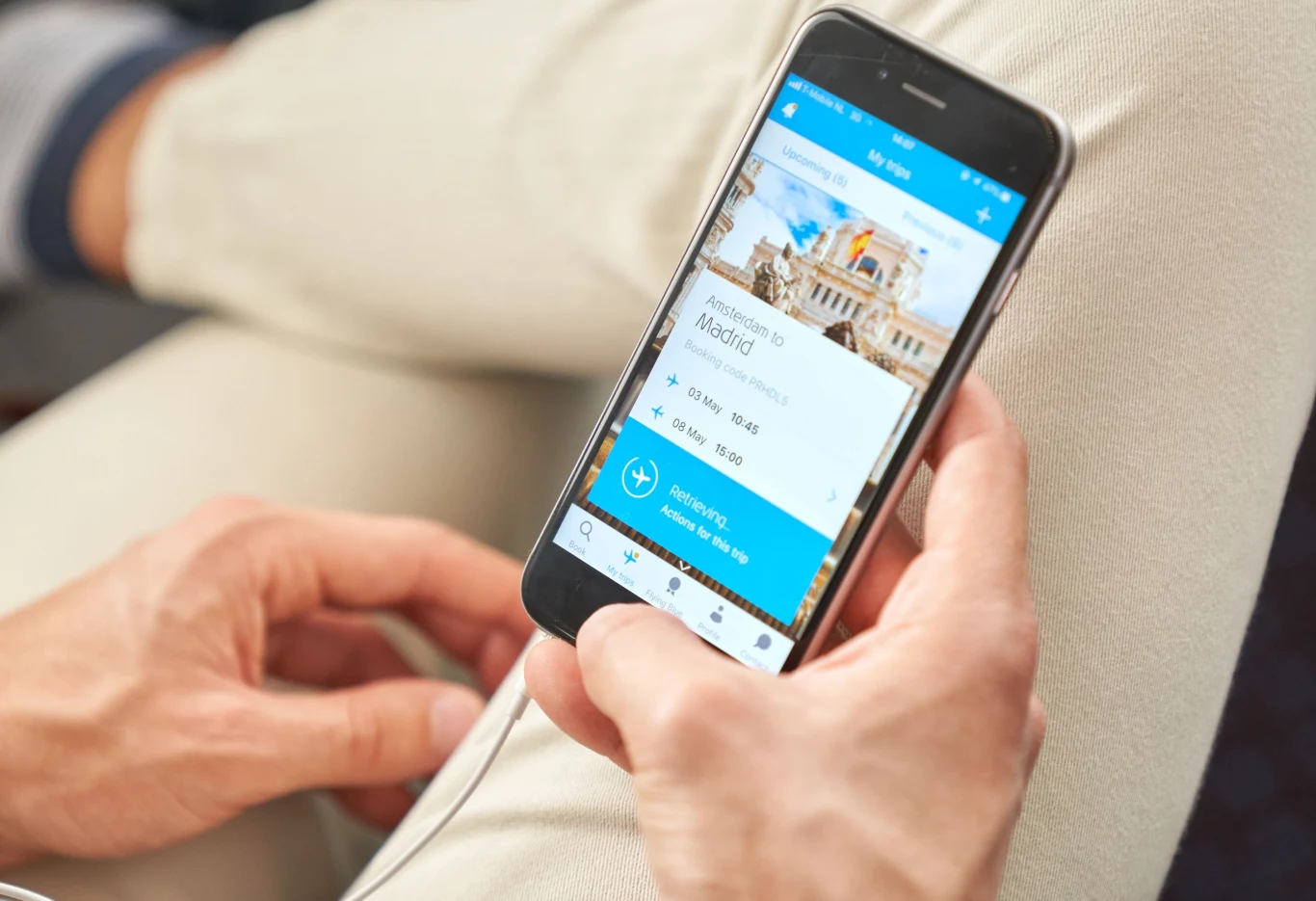 KLM włącza darmowy internet dla pasażerów. Osoba trzymająca smartfon z otwartą aplikacją podróżniczą na ekranie, planująca podróż do Madrytu, widoczne szczegóły lotu oraz fragmenty ubrań użytkownika.