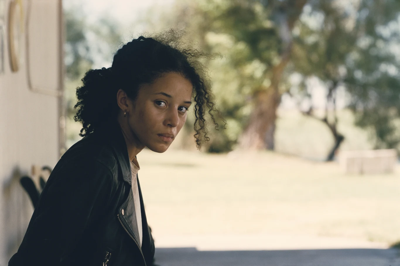 Młoda kobieta z ciemnymi kręconymi włosami ubrana w czarną kurtkę, siedzi na zewnątrz, patrząc w stronę aparatu. W tle rozmyte drzewa i światło dzienne.