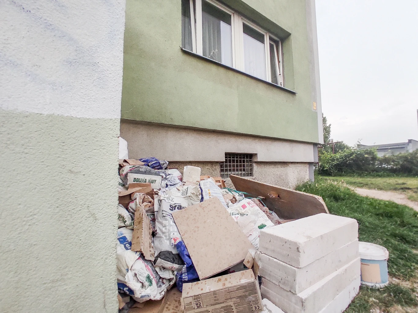 Odpady remontowe należy wywieźć do PSZOK. Sterta odpadów budowlanych, worków z zaprawą, kartonów i styropianowych bloczków zgromadzona pod ścianą zielonego budynku mieszkalnego, częściowo blokująca okno piwniczne, w tle fragment zieleni i ścieżki.