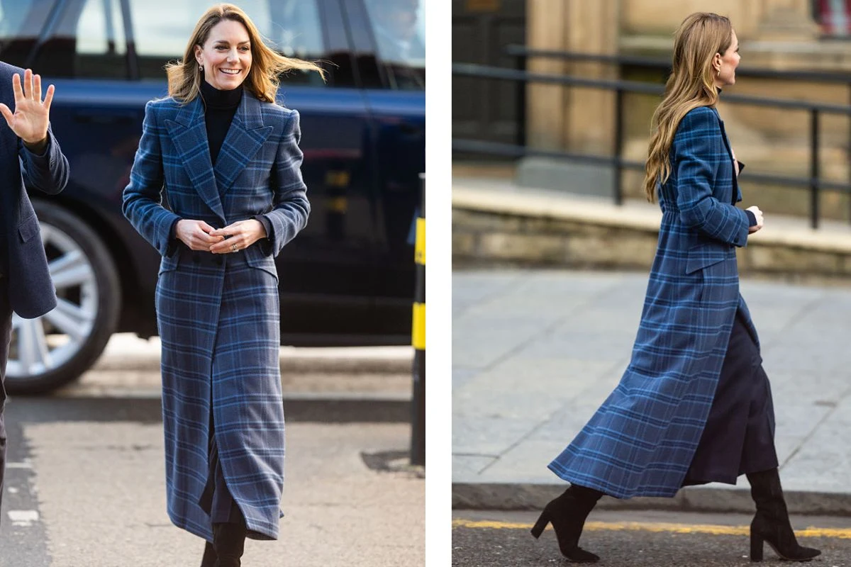 Kobieta w długim, granatowym płaszczu w kratę, czarnych butach na obcasie i czarnym golfie spaceruje po ulicy. W tle widoczne są częściowo samochód terenowy oraz fragment miejskiego chodnika i schodów.