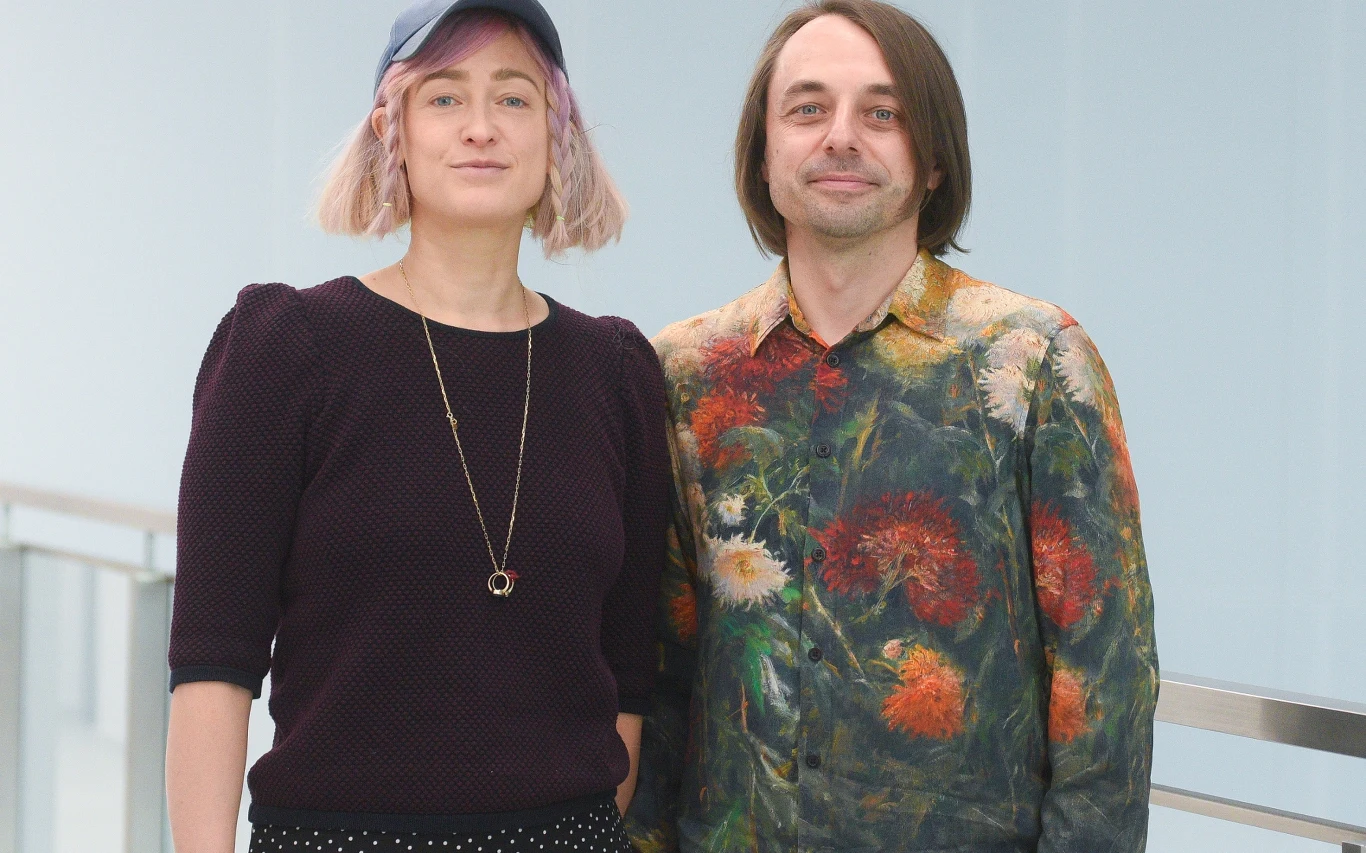 Matylda Damięcka i Radosław Łukasiewicz Dwie osoby stoją obok siebie w jasnym, nowoczesnym wnętrzu. Kobieta ma różowe włosy, ciemną bluzkę z długim rękawem i naszyjnik, mężczyzna nosi kolorową koszulę z motywem kwiatowym. Oboje patrzą wprost w obiektyw i wyglądają na zrelaksowanych.