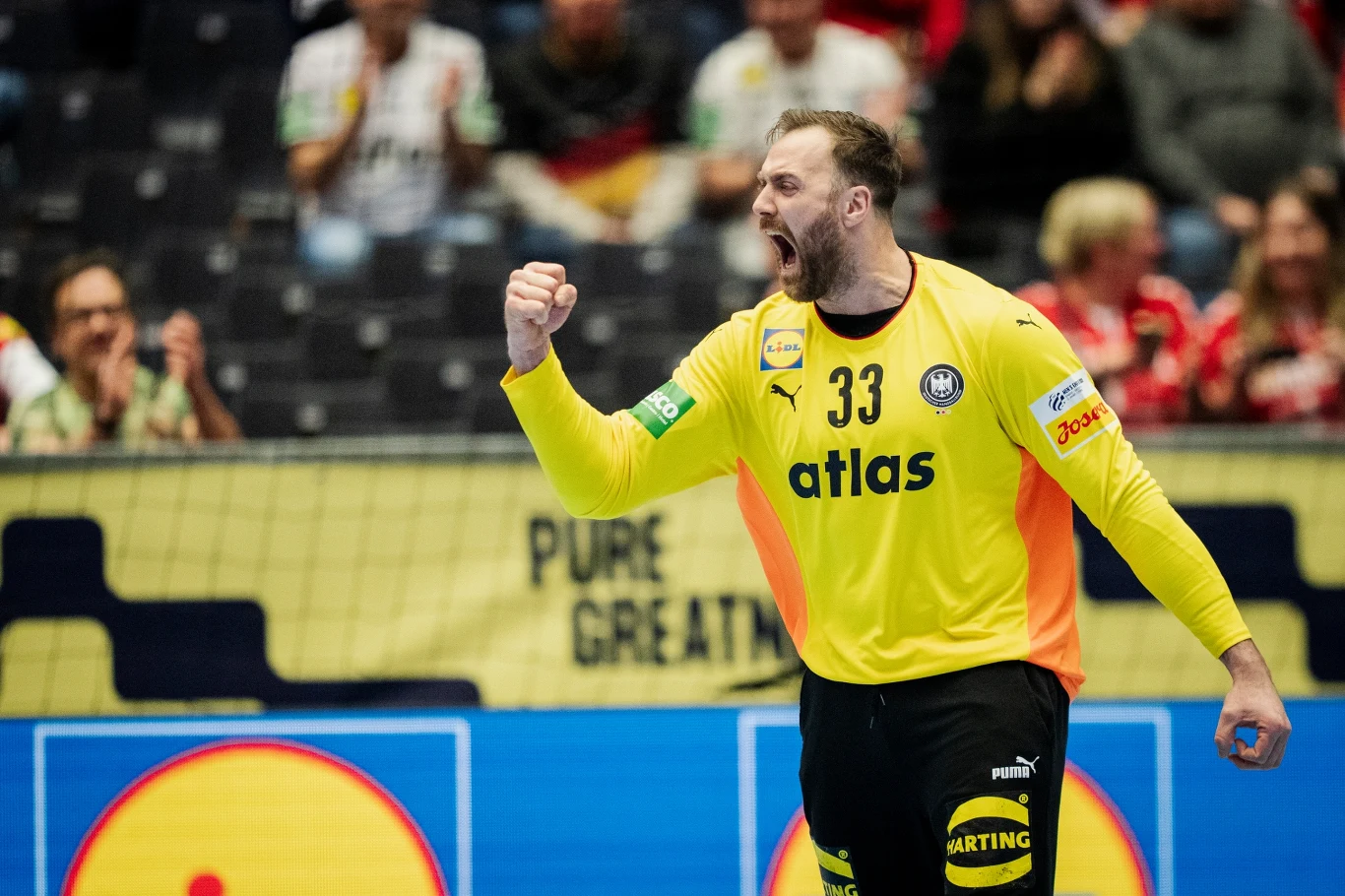 Andreas Wolff A handball player in a yellow goalkeeper uniform with a raised, clenched fist expresses strong emotions, likely joy after a triumphant play, with the audience and advertisements visible in the background.
