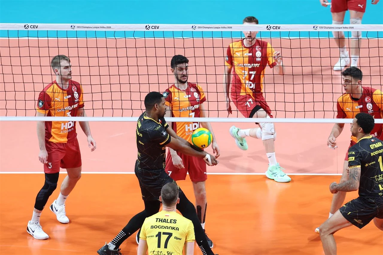 Zawodnicy drużyn siatkarskich w trakcie rozgrywki przy siatce, jeden z graczy w żółto-czarnym stroju odbija piłkę, wokół kilku przeciwników w czerwono-pomarańczowych strojach, dynamiczna akcja na boisku.