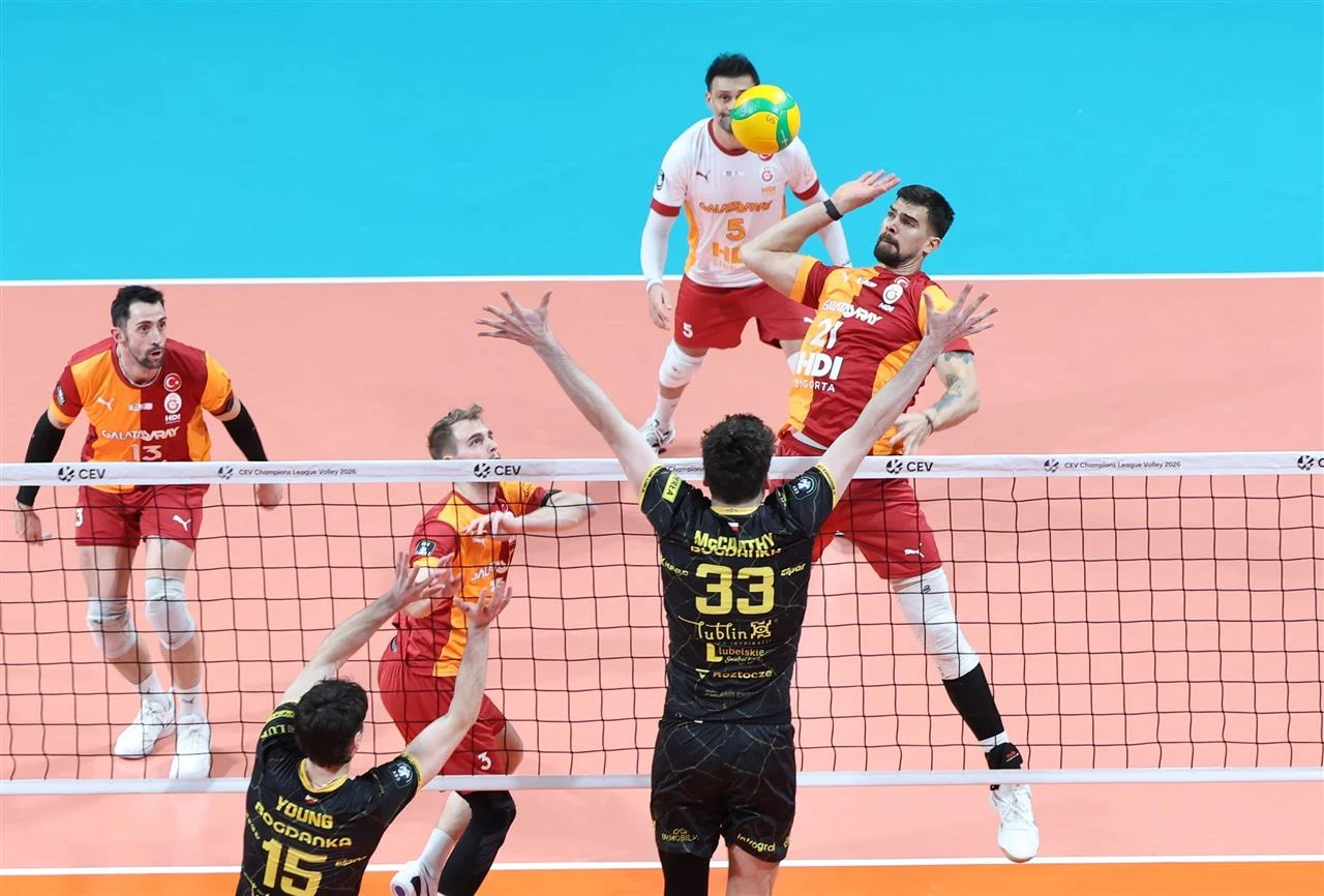 Dwóch zawodników siatkarskich w powietrzu po obu stronach siatki, jeden wykonuje atak, drugi próbuje blokować. W tle widocznych jest kilku innych graczy i fragment boiska. Ujęcie dynamiczne, podkreślające rywalizację sportową.