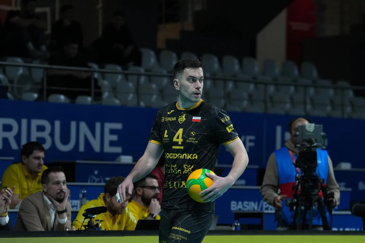 Zawodnik siatkarski w czarnym stroju z numerem 4 trzyma kolorową piłkę do siatkówki. W tle sędziowie oraz operator kamery, trybuny częściowo wypełnione widzami, dominuje sportowa i profesjonalna atmosfera hali.