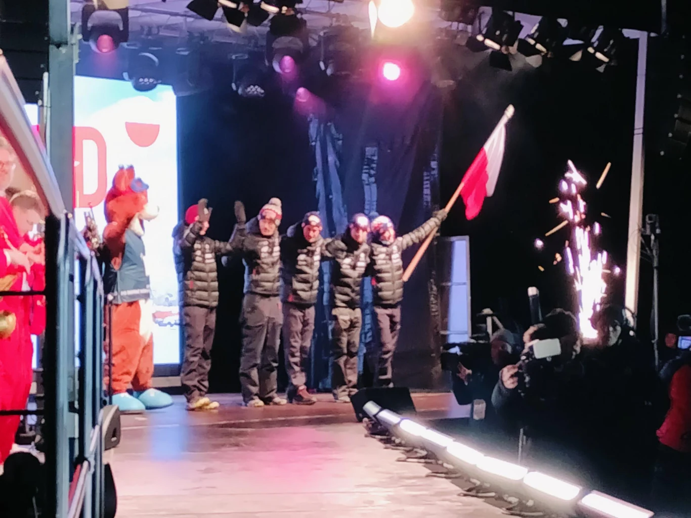 Grupa osób ubranych w kurtki zimowe stoi na scenie, jedna z nich trzyma polską flagę, na scenie obecna maskotka w kostiumie oraz widoczne efekty świetlne i pirotechniczne, wokół zebrani są fotoreporterzy i obserwatorzy.