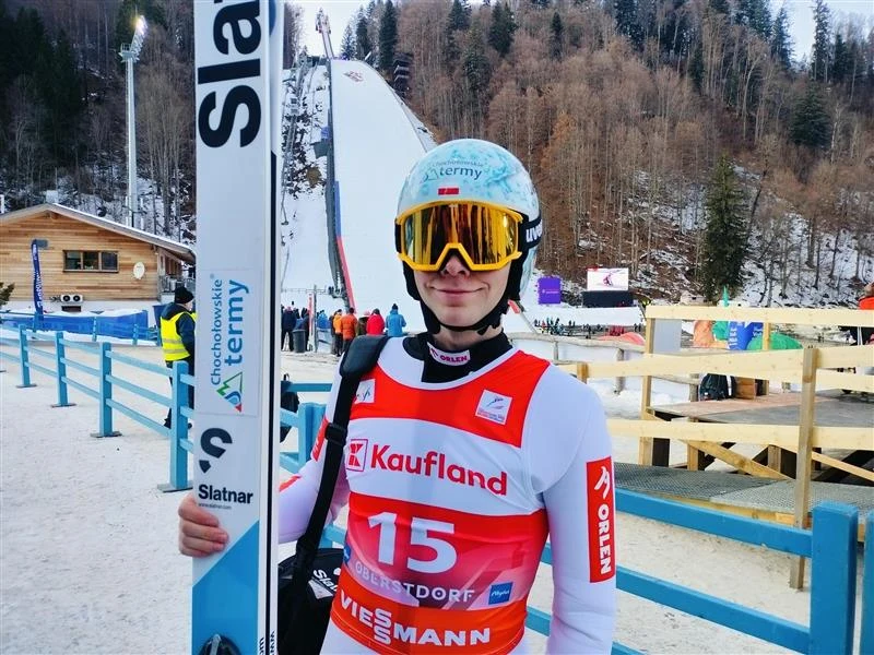 Skoczek narciarski ubrany w kombinezon startowy i kask, trzymający narty na tle zimowego obiektu sportowego z widoczną skocznią narciarską oraz budynkami i drzewami w tle.