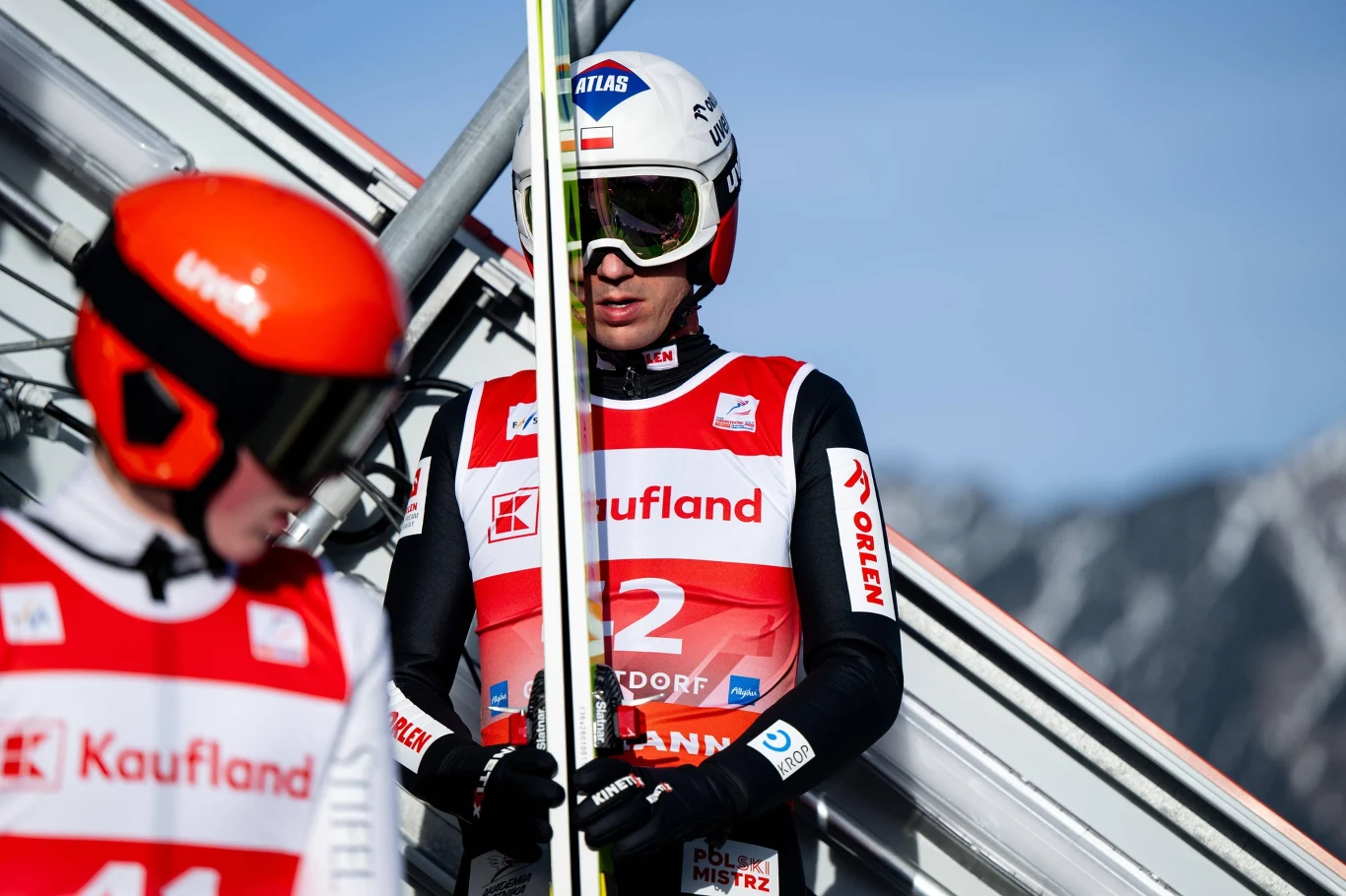 Dwóch skoczków narciarskich w strojach startowych i kaskach, jeden trzyma narty i stoi na platformie skoczni, w tle widoczne góry.