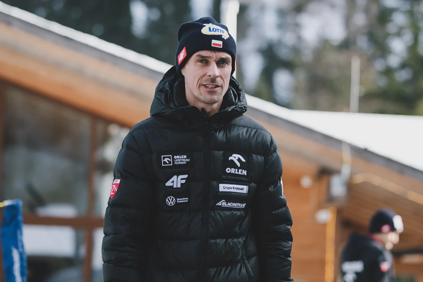 Mężczyzna w sportowej kurtce z licznymi logotypami sponsorów oraz czapce z daszkiem spaceruje na świeżym powietrzu, w tle rozmyty budynek oraz druga osoba.