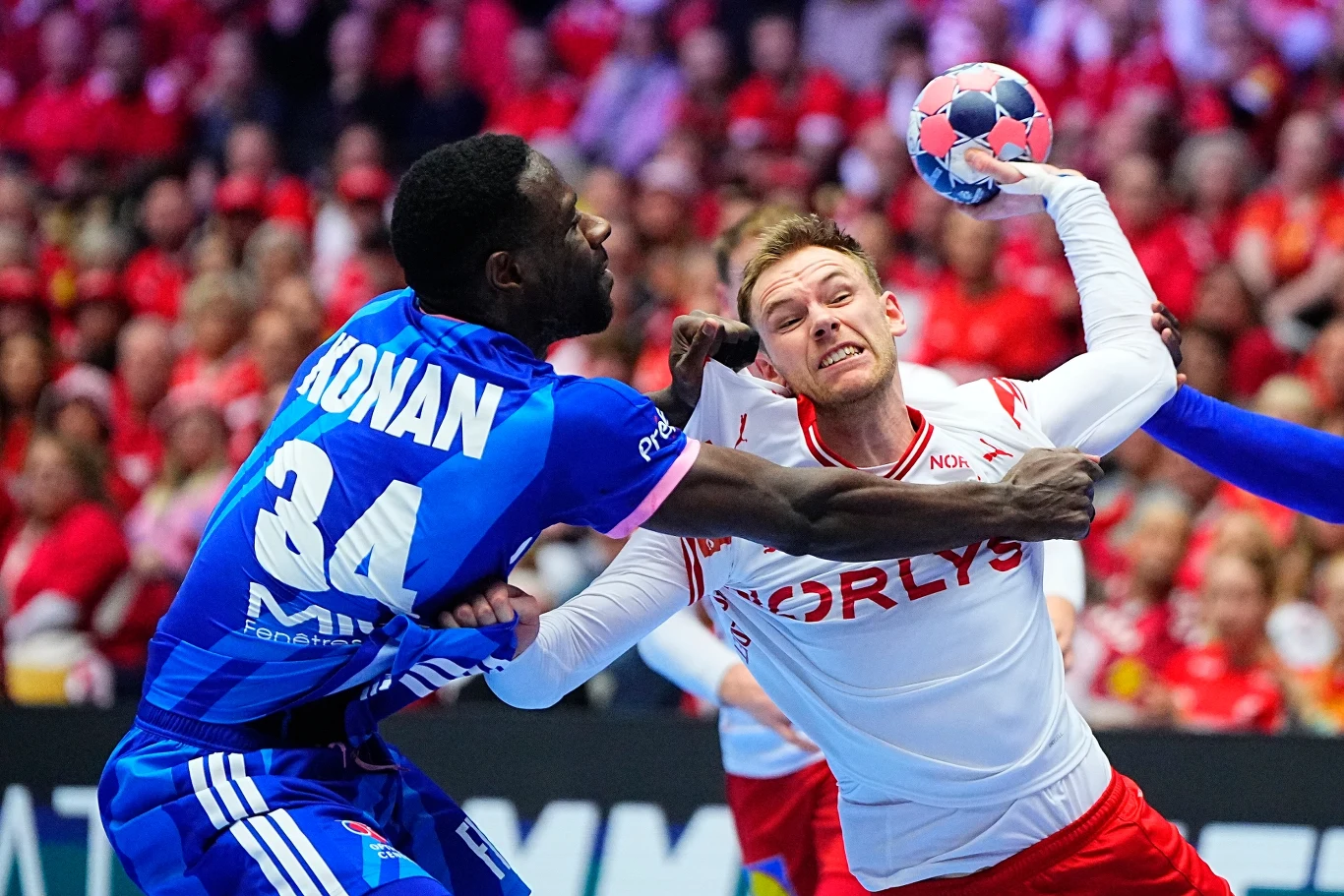 Dwóch zawodników podczas dynamicznej walki o piłkę ręczną, jeden ubrany w niebieski strój z numerem 34, drugi w biało-czerwonym stroju, obydwaj zaangażowani fizycznie, a w tle kibice na trybunach.