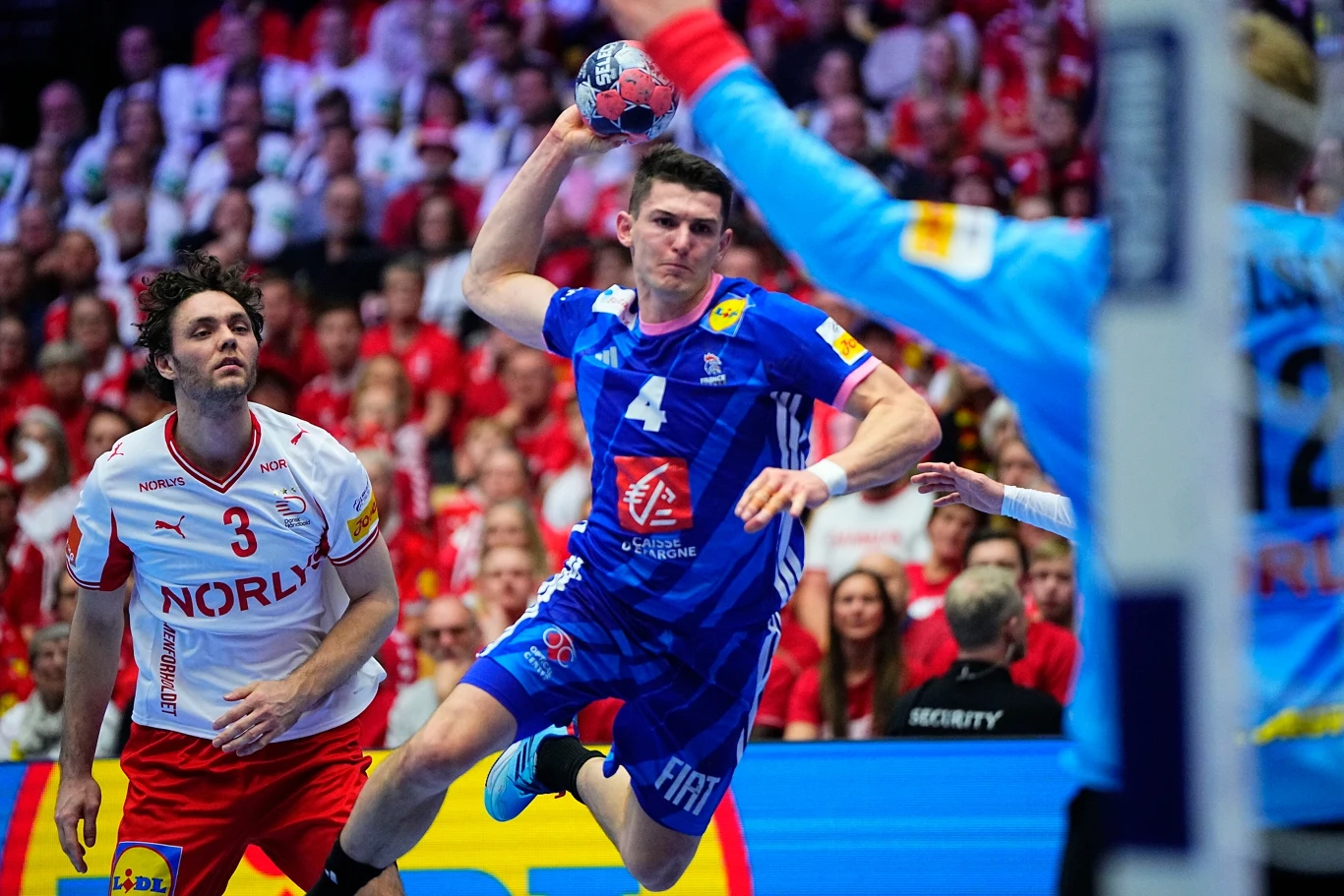 Zawodnik w niebieskim stroju wyskakuje do rzutu podczas meczu piłki ręcznej, próbując zdobyć bramkę. W tle widoczni pozostali zawodnicy drużyny przeciwnej w białych strojach oraz kibice na trybunach.