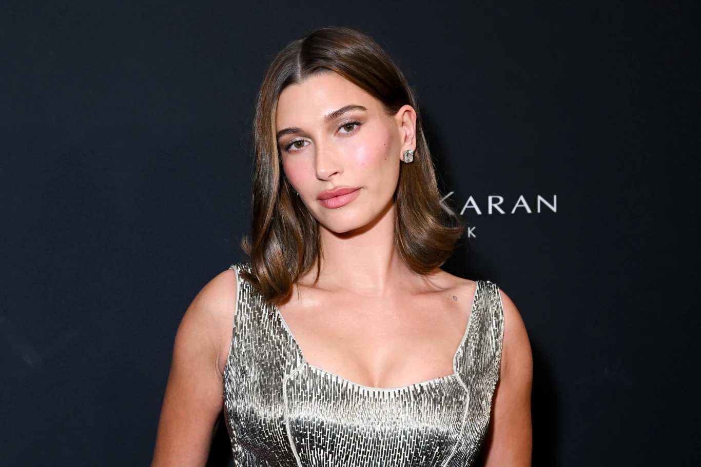 Kobieta o jasnej karnacji i brązowych włosach do ramion pozuje na tle ciemnego tła ubrana w srebrną, błyszczącą sukienkę bez rękawów, delikatny makijaż oraz subtelna biżuteria podkreślają elegancki wygląd.