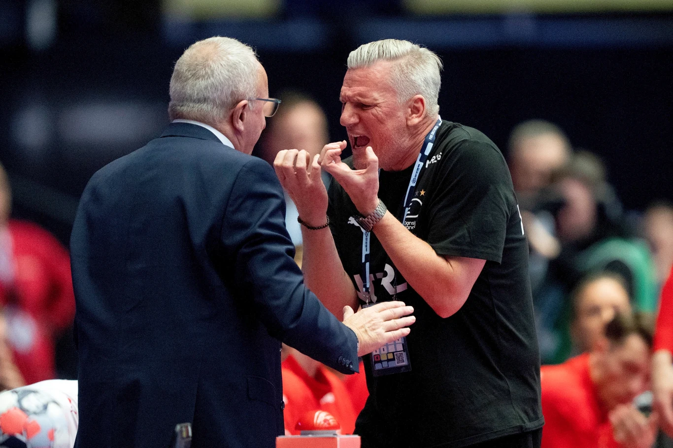 Dwóch mężczyzn prowadzi intensywną rozmowę, jeden energicznie gestykuluje, drugi słucha uważnie. W tle widoczni są kibice oraz sportowe otoczenie.