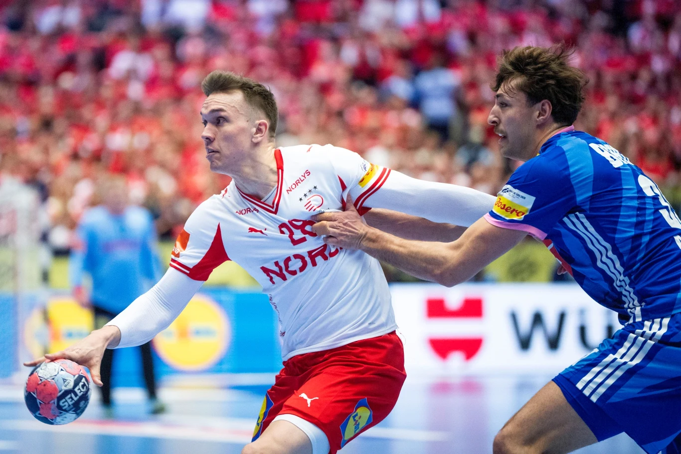 Dwóch zawodników podczas dynamicznej akcji na meczu piłki ręcznej, jeden z nich w białoczerwonym stroju z napisem 'Nor', druga osoba w niebieskiej koszulce próbuje go zatrzymać chwytając za koszulkę, w tle trybuny pełne kibiców.