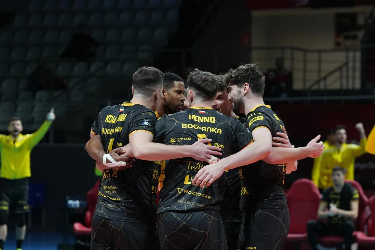 Grupa siatkarzy drużyny Bogdanka LUK Lublin ubrana w czarne stroje sportowe z żółtymi napisami, trzyma się w ścisłym kręgu na boisku podczas meczu sportowego, w tle widać innych zawodników i puste trybuny.