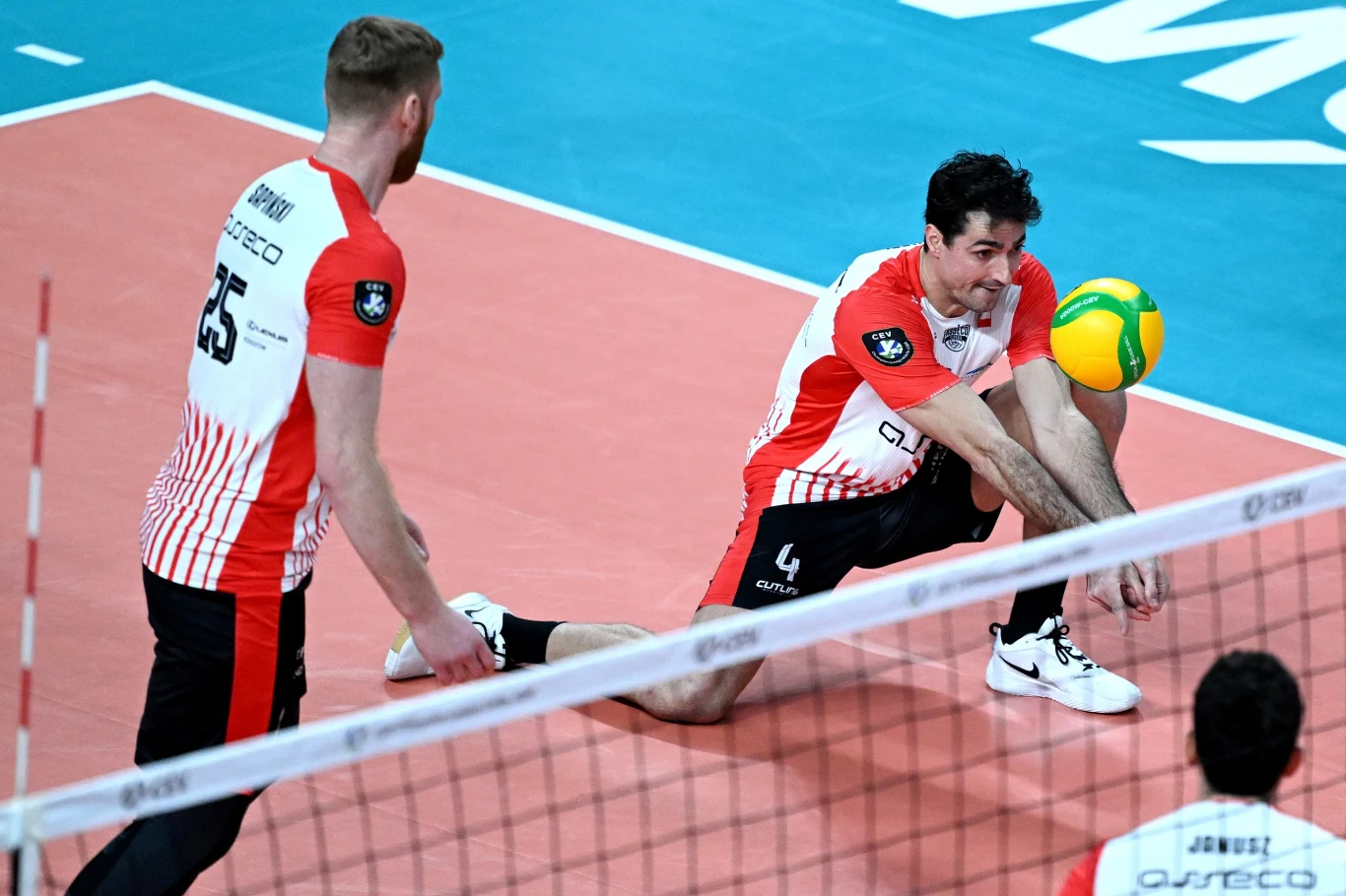 Dwóch siatkarzy ubranych w biało-czerwone stroje drużynowe podczas dynamicznej akcji na boisku, jeden z nich wykonuje odbiór piłki, drugi obserwuje sytuację. W tle widoczna linia siatki oraz fragment boiska.