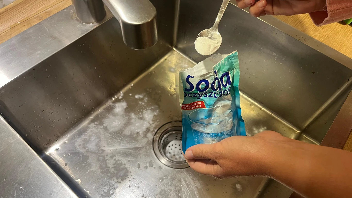 Ręka wsypuje sodę oczyszczoną z opakowania do zlewu kuchennego, wokół widoczny osad sody na powierzchni zlewu ze stali nierdzewnej.