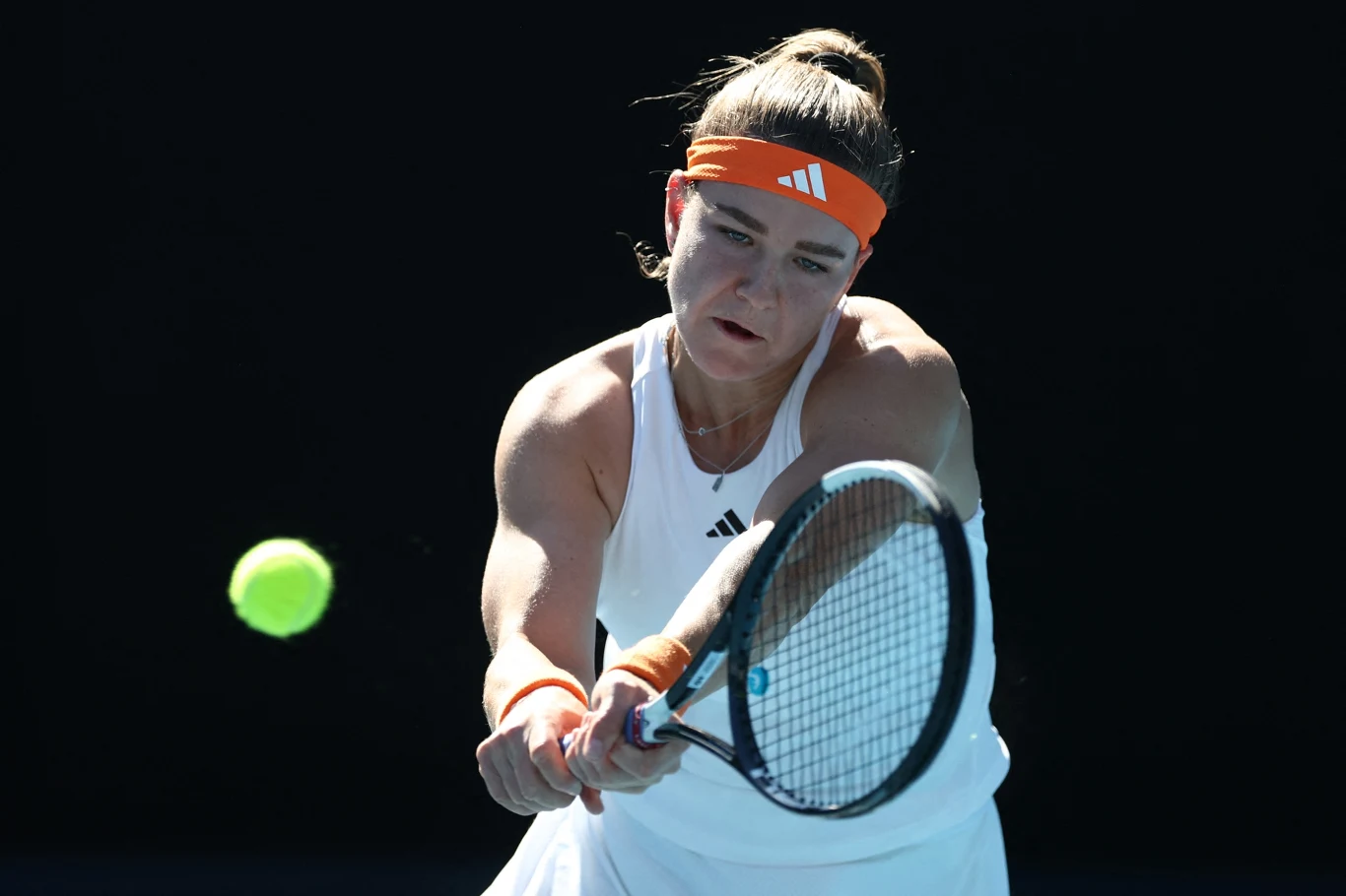 Kobieta w białym stroju sportowym z pomarańczową opaską na głowie uderza dwuręcznym backhandem żółtą piłkę tenisową, skupiona na grze.