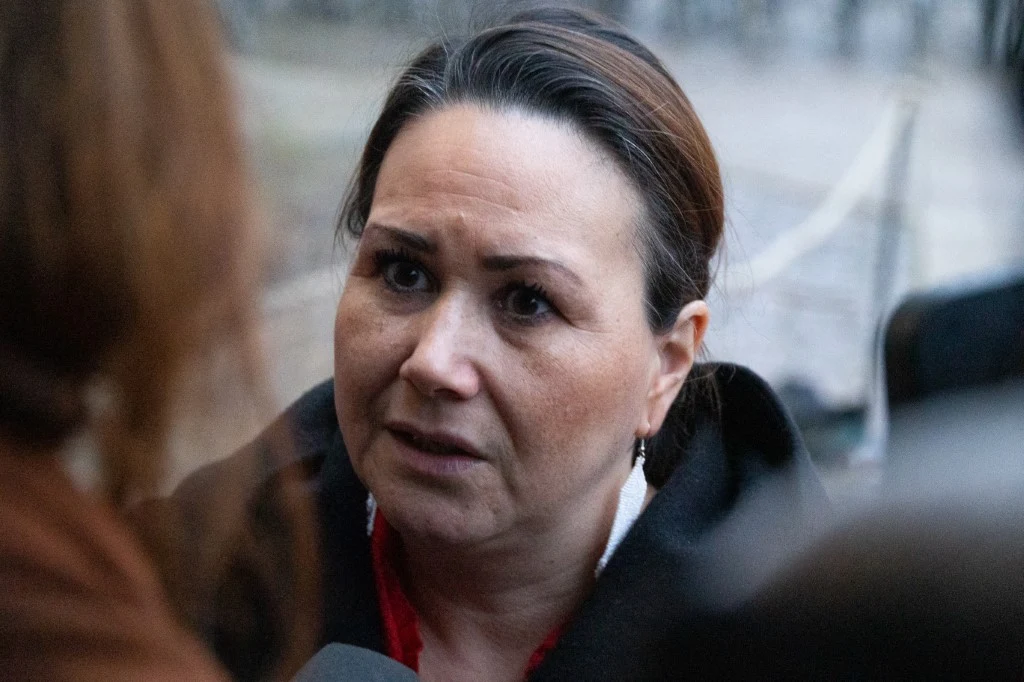 Kobieta o ciemnych włosach spiętych do tyłu patrzy z powagą na rozmówcę, stojąc na zewnątrz w zimnej scenerii. Zbliżenie na twarz, lekko zaniepokojony wyraz twarzy.