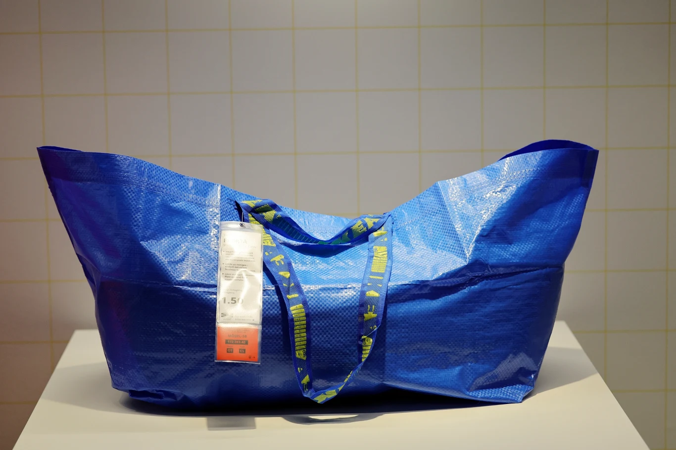 IKEA FRAKTA Duża niebieska torba zakupowa IKEA z metką, leżąca na jasnym podłożu na tle białej ściany o kratkowanej strukturze.