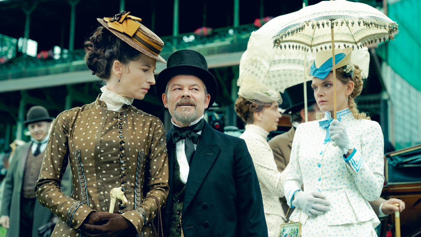 Trzy osoby ubrane w stroje z końca XIX wieku, kobieta w brązowej sukni i kapeluszu patrzy na mężczyznę w cylindrze i białej muszce, obok stoi druga kobieta w jasnym stroju z parasolką chroniącą przed słońcem. W tle widoczne są trybuny oraz inne osoby r...