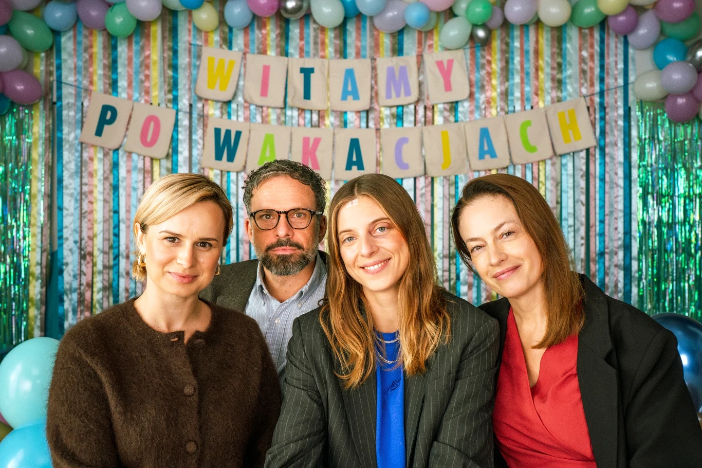 Cztery osoby uśmiechające się do aparatu, w tle kolorowe balony, girlanda oraz napis 'Witamy po wakacjach', wszystko utrzymane w radosnej, powitalnej atmosferze.