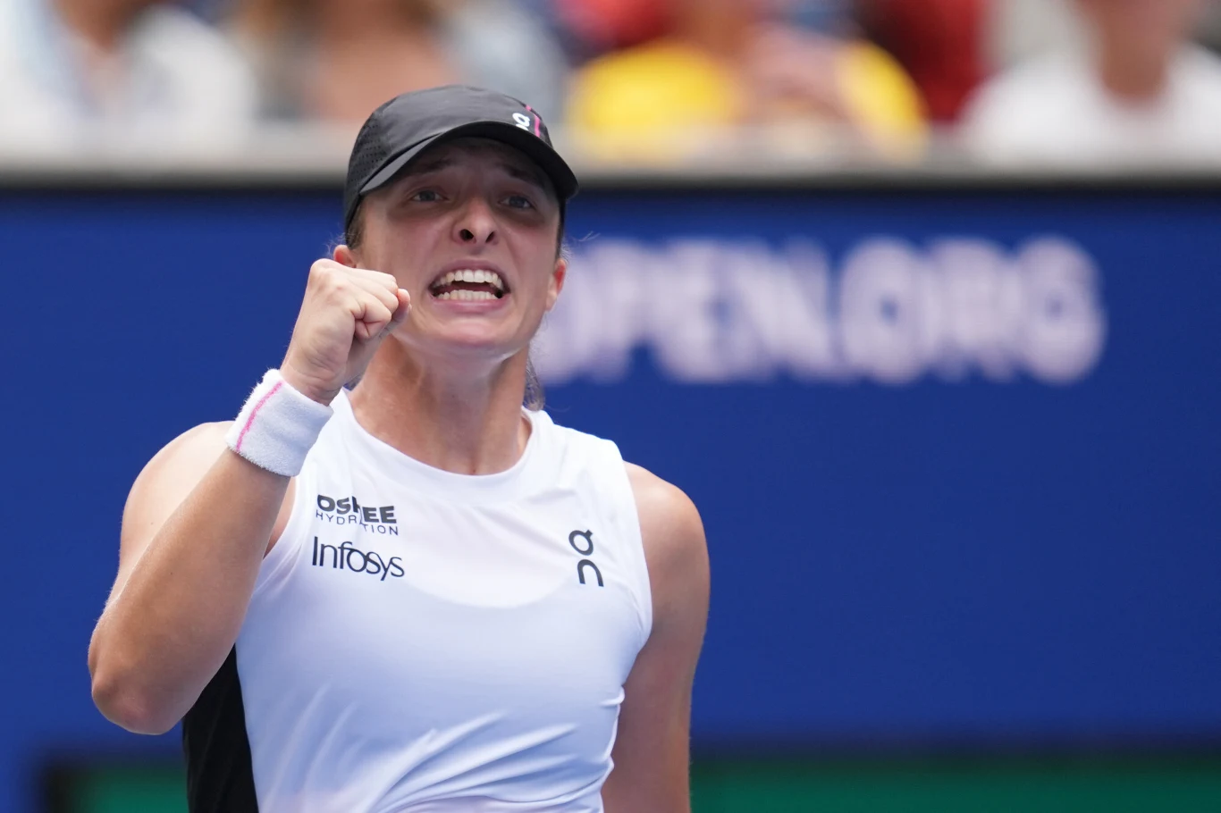 Tenisistka w sportowym stroju unosi zaciśniętą pięść w geście triumfu, wyrażając silne emocje podczas meczu na korcie tenisowym, na tle napisu OPEN.