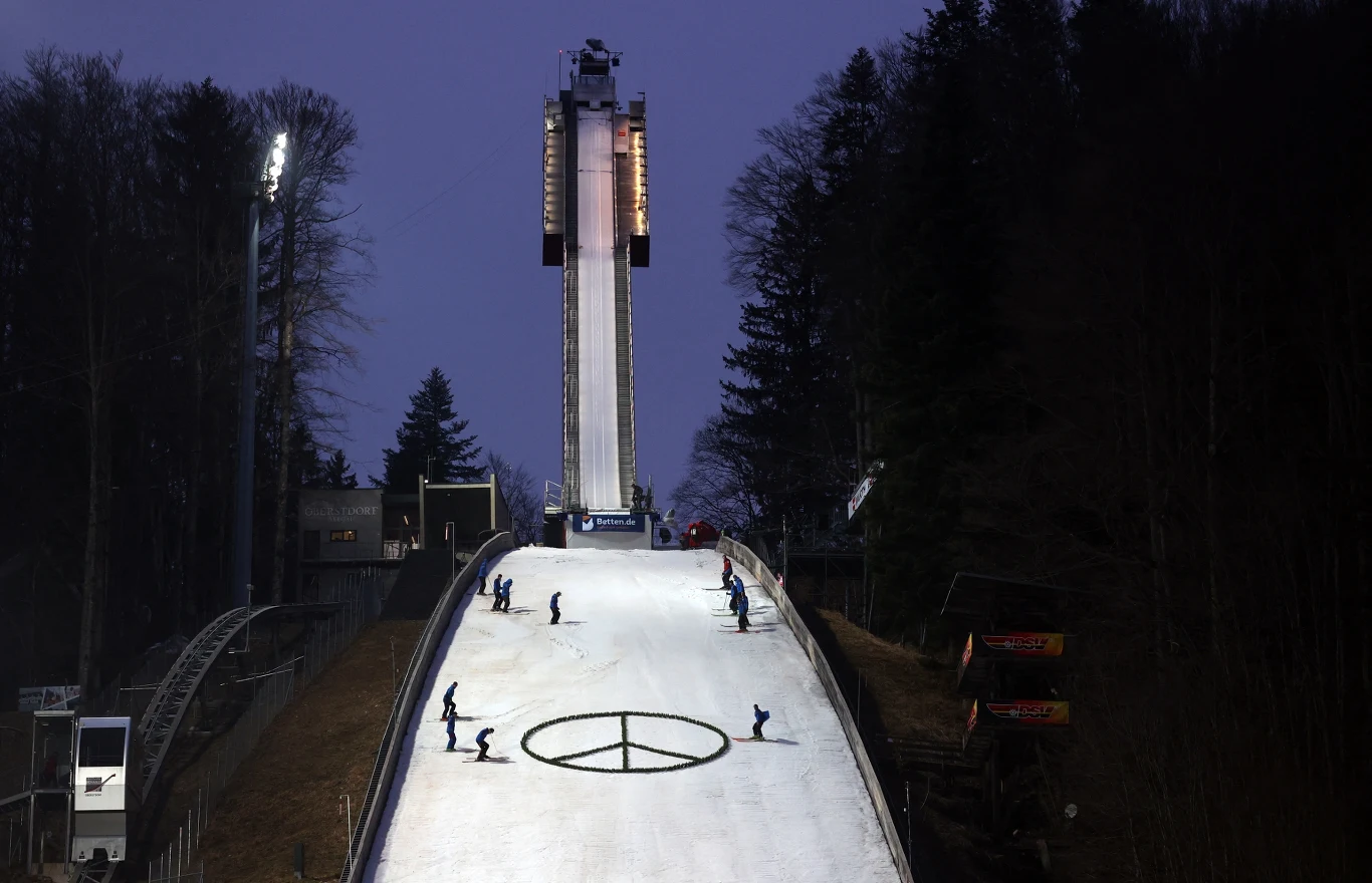 Oświetlona skocznia narciarska po zmroku, na której znajduje się duży symbol pokoju wykonany na śniegu, kilku pracowników przygotowuje tor zjazdu, a w tle widoczne są zalesione wzgórza i wysokie światła oświetleniowe.