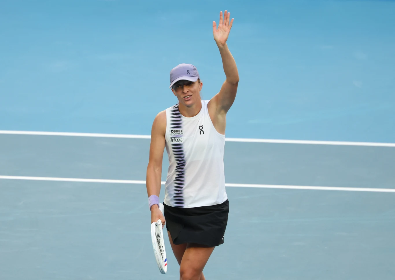 Tenisistka na korcie w sportowym stroju unosi rękę w geście powitania lub podziękowania, w drugiej dłoni trzyma rakietę tenisową, jasnoniebieskie tło boiska.