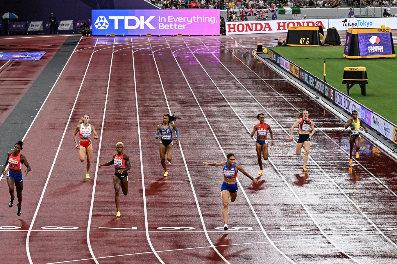 Finisz biegu lekkoatletycznego kobiet na stadionie podczas deszczu, zawodniczki z różnych krajów walczące o osiągnięcie mety, widoczne mokre bieżnie i reklamy wokół toru.