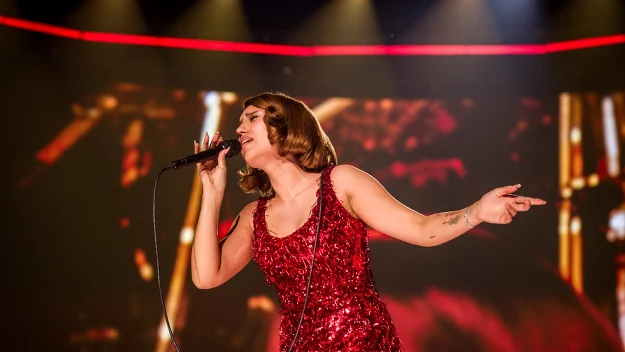 Kobieta w czerwonej, błyszczącej sukni trzyma mikrofon i śpiewa na scenie koncertowej, podświetlona jasnym światłem oraz otoczona dynamicznym tłem wizualizacji.