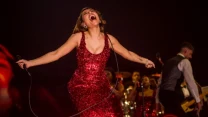 Wokalistka w cekinowej czerwonej sukni śpiewa na scenie, trzymając mikrofon, bije energią i ekspresją, w tle zespół muzyczny oraz intensywne czerwone światła sceniczne.
