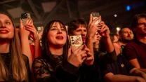 Grupa młodych ludzi w tłumie na koncercie, uśmiechnięci, trzymają telefony komórkowe i nagrywają występ ze sceny, w tle rozświetlone światłami wnętrze hali.
