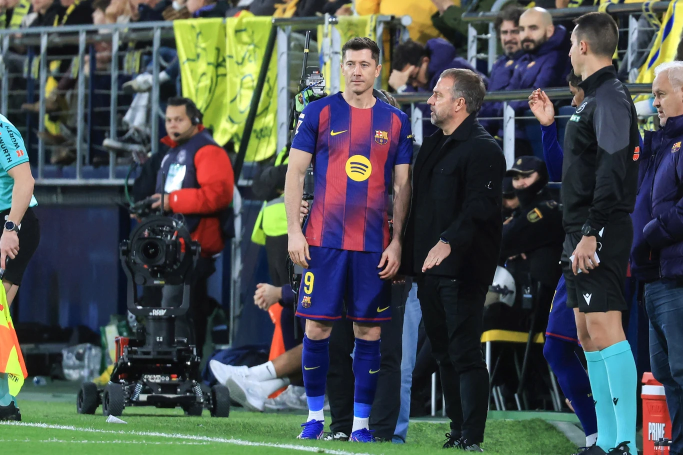 Piłkarz w stroju FC Barcelona stoi przy linii bocznej boiska, obok niego trener oraz sędzia, w tle widoczna publiczność i barierki stadionowe.