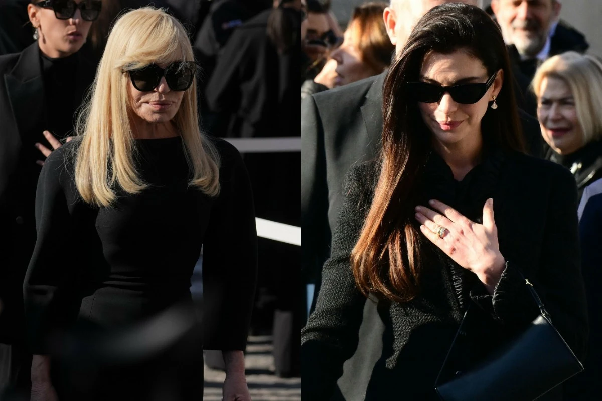 Dwie elegancko ubrane kobiety w czerni stoją obok siebie na tle innych osób, obie mają okulary przeciwsłoneczne, jedna z nich ma blond włosy, druga długie, ciemne włosy i trzyma dłoń na piersi.