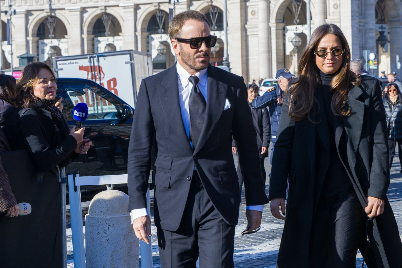 Elegancko ubrany mężczyzna w garniturze i kobieta w ciemnym płaszczu idą razem w przestrzeni miejskiej wśród innych ludzi, w tle monumentalna architektura oraz reporterzy z kamerą.