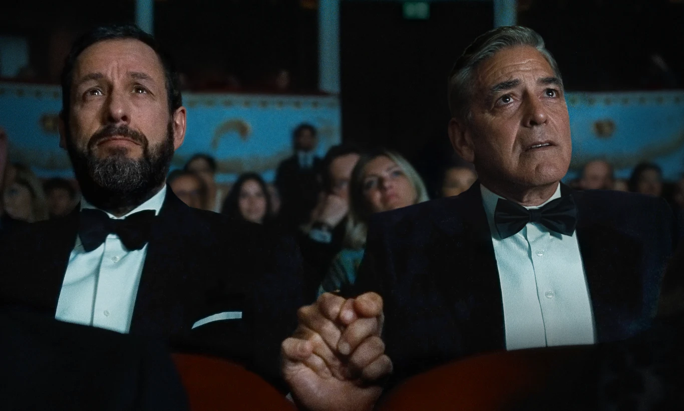 Dwaj mężczyźni w smokingu siedzą obok siebie na widowni, trzymając się za ręce i patrząc w jednym kierunku z poważnym wyrazem twarzy, w tle widoczne inne osoby.