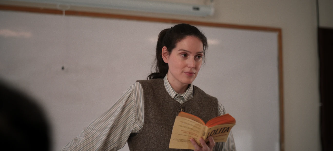 Młoda kobieta stojąca przed tablicą, ubrana w koszulę i kamizelkę, trzyma otwartą książkę, skupiając uwagę na rozmówcy.