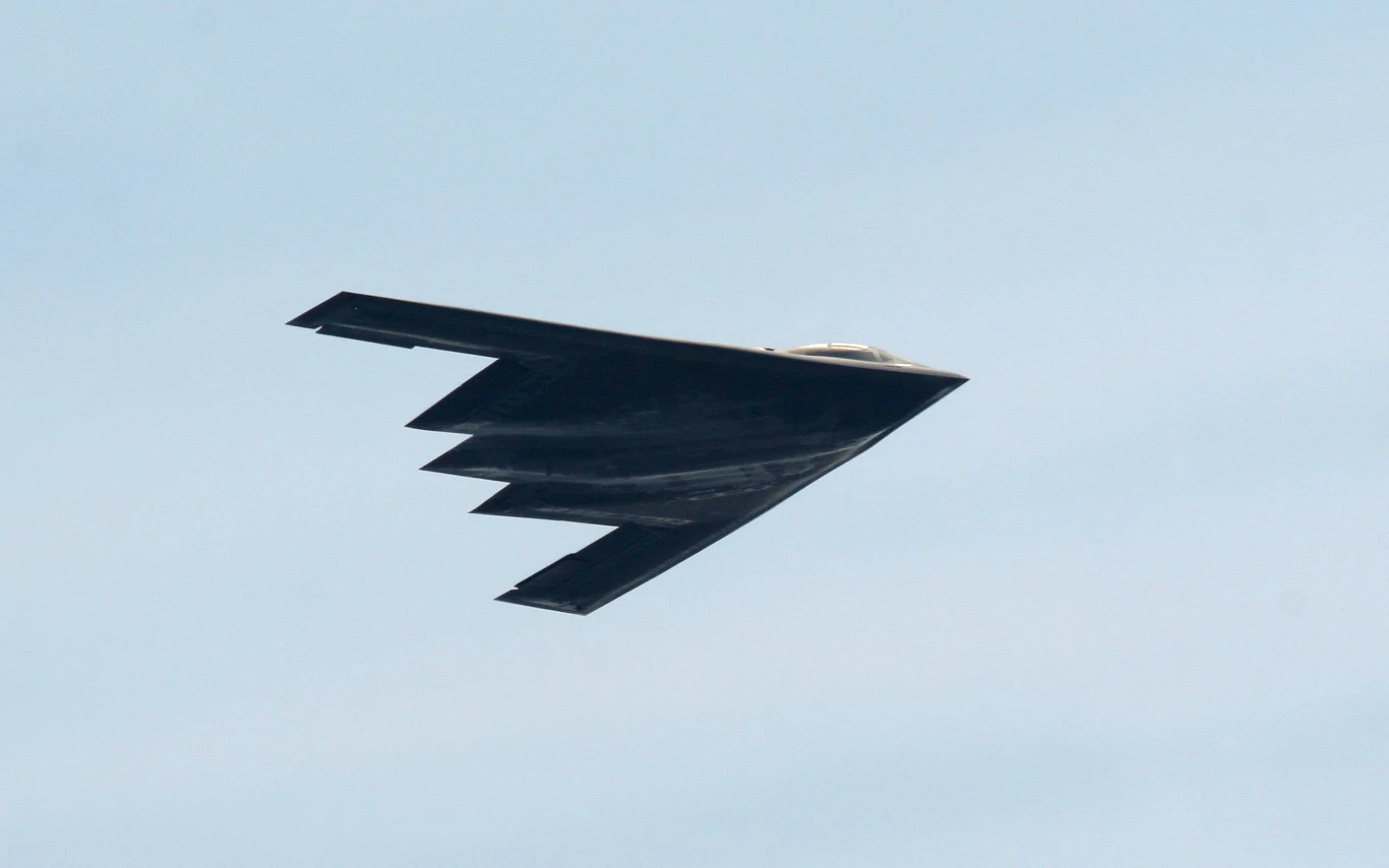 Bombowiec B-2 Spirit Czarny bombowiec stealth o charakterystycznym kształcie latający na tle jasnego nieba.
