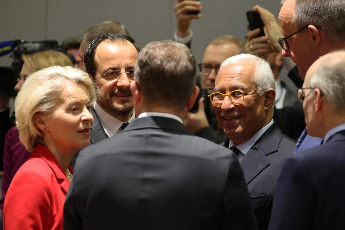 Grupa elegancko ubranych osób, prawdopodobnie polityków lub urzędników wysokiego szczebla, rozmawia ze sobą podczas oficjalnego spotkania. W tle widoczni są inni uczestnicy oraz osoby robiące zdjęcia telefonami.