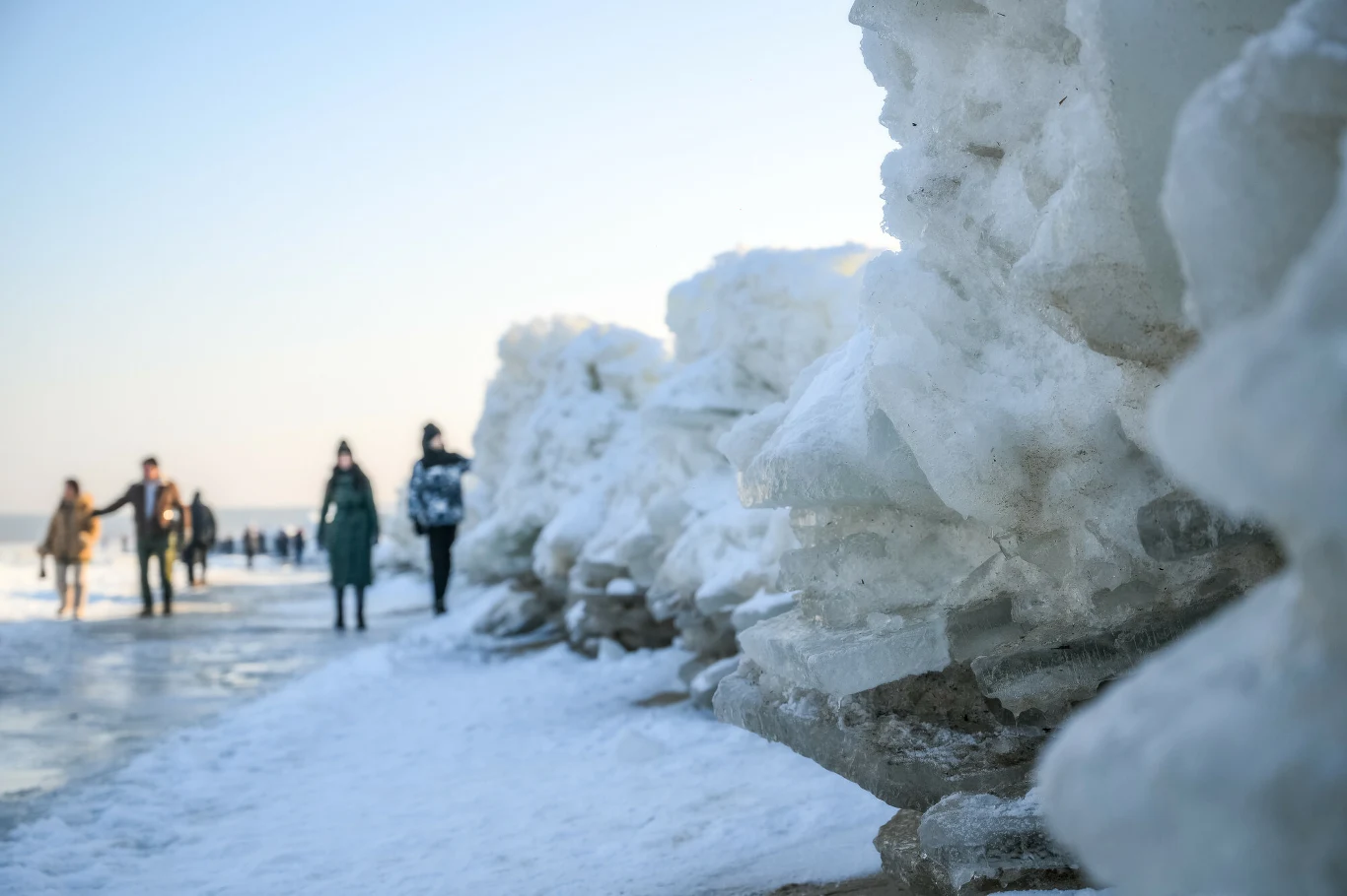 Lodowe kryształy tworzące wysokie zaspy nad brzegiem zamarzniętego zbiornika wodnego, na którym spacerują ludzie ubrani w zimowe płaszcze. W tle widać rozmyte sylwetki kolejnych osób.