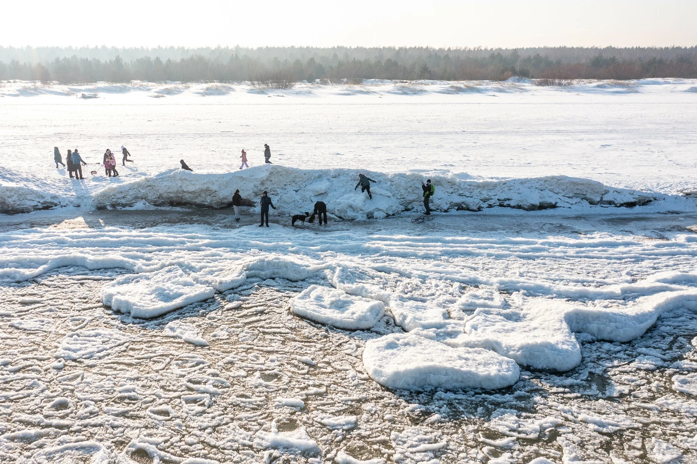 Grupa ludzi spaceruje oraz bawi się na zamarzniętej rzece pokrytej grubą warstwą śniegu i lodu. W tle widoczny jest las oraz otwarta, zimowa przestrzeń. Duże kry lodowe tworzą nieregularne formacje na powierzchni wody.