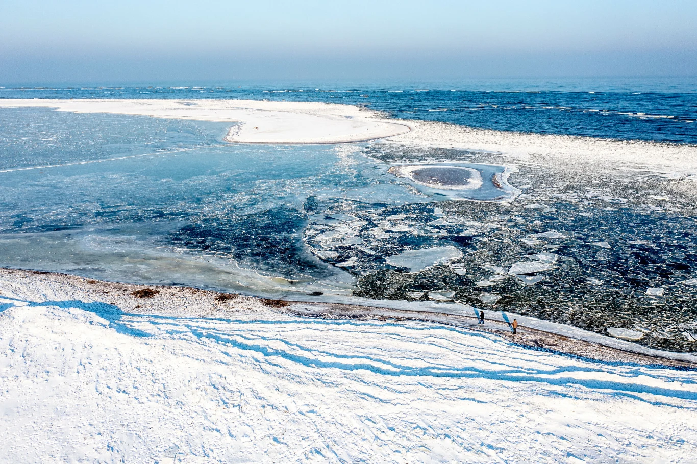 Zimowy krajobraz nadmorski z pokrywą lodową, widocznym brzegiem pokrytym śniegiem oraz zamarzniętym fragmentem morza z oddzielającą się wyspą lodową. Na śnieżnej ścieżce maszerują dwie osoby.