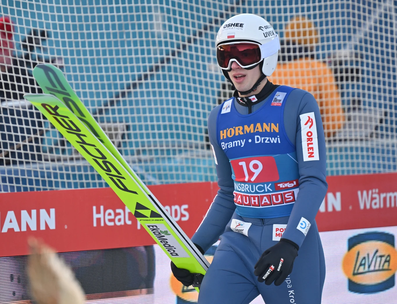 Skoczek narciarski w stroju sportowym i kasku, trzymający narty przy siatce ochronnej na tle banerów sponsorów