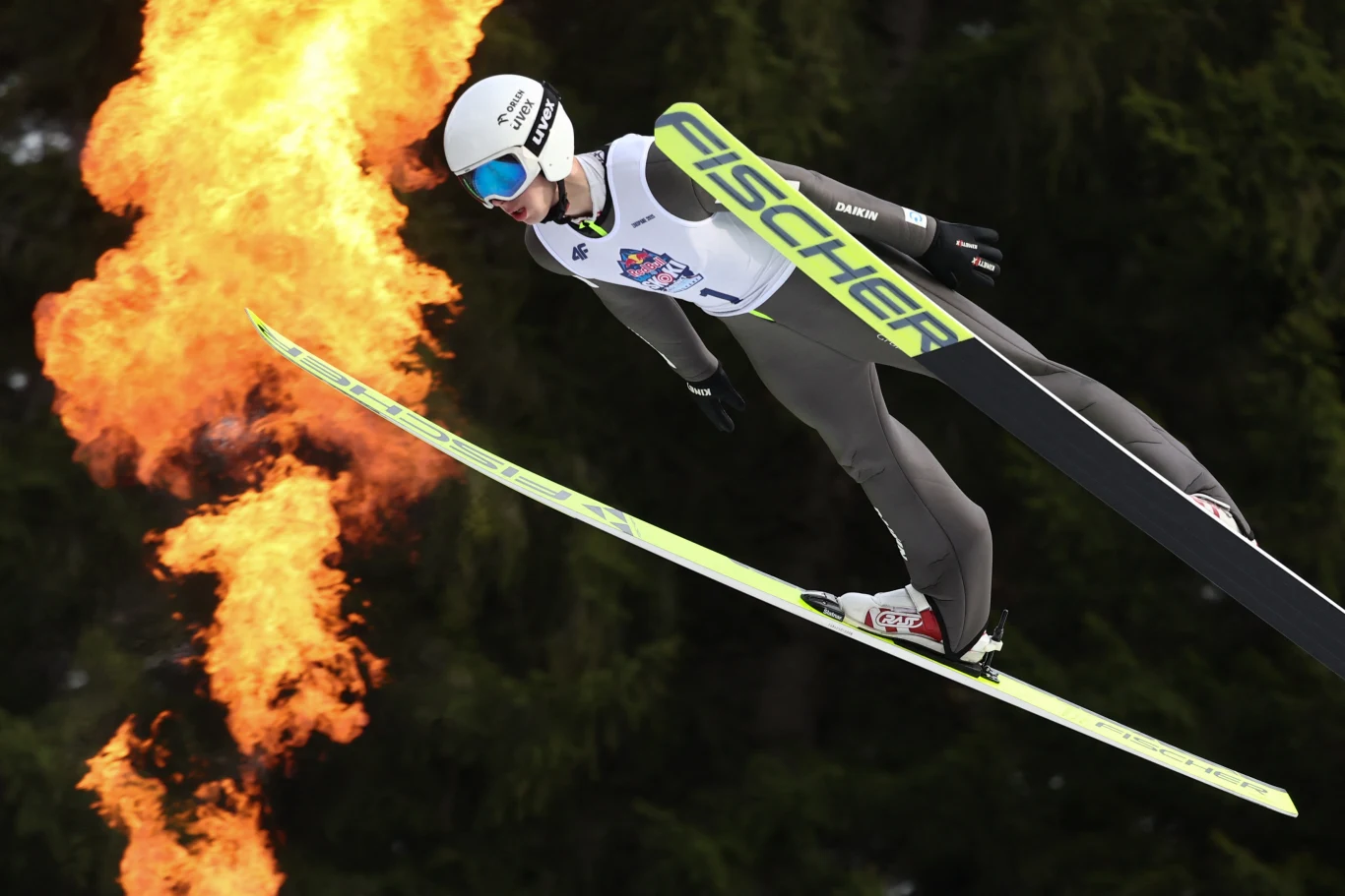 Skoczek narciarski w locie z płomieniem ognia w tle, na tle ciemnego lasu, w profesjonalnym stroju oraz kasku, lecący na nartach marki Fischer.