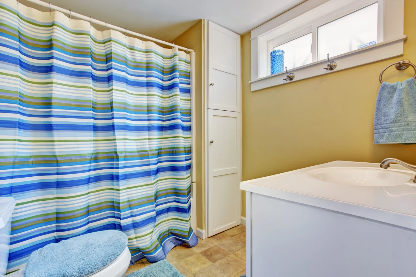 Łazienka z jasnobrązowymi ścianami, kolorową prysznicową zasłoną w niebiesko-zielone pasy, białymi meblami, umywalką i ręcznikiem na wieszaku obok okna.