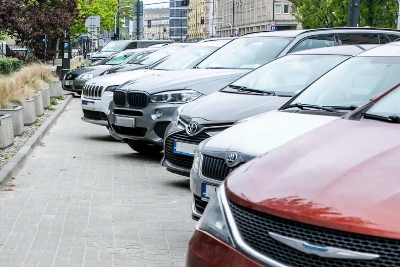 Kierowcy stracą tysiące miejsc do parkowania. Duże zmiany w stolicy Szereg zaparkowanych samochodów różnych marek ustawionych pod kątem przy krawędzi chodnika w centrum miasta, w tle niska roślinność oraz zabudowania miejskie.