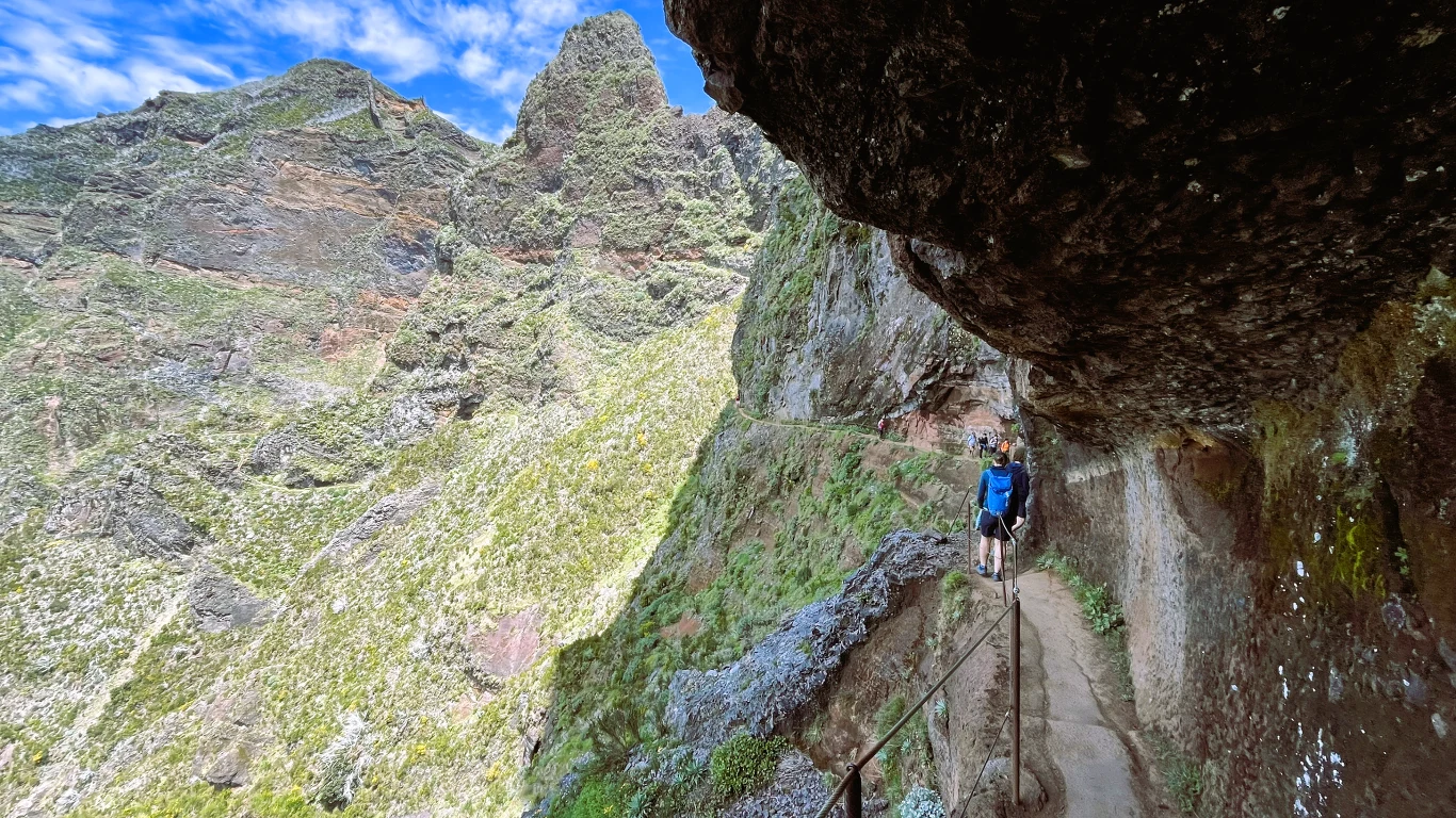 Wąska, wykuta w skale ścieżka prowadzi wzdłuż urwiska, z jednej strony zabezpieczona metalową barierką, otoczona imponującym górskim krajobrazem, pod niebieskim niebem z umiarkowaną ilością chmur; samotny turysta idzie szlakiem.