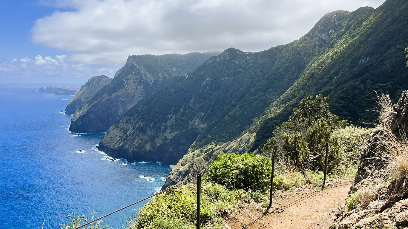 Widok skalistego wybrzeża z wysokimi klifami porośniętymi zielenią, nad którym rozciąga się błękitne morze, częściowo przykryte chmurami niebo i ścieżka prowadząca wzdłuż krawędzi urwiska.
